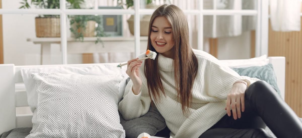 Smiling young lady in warm knitted sweater enjoying fresh sushi on wicker tray while relaxing on cozy sofa with soft cushions in modern living room