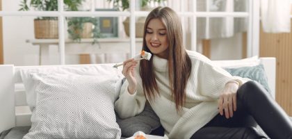 Smiling young lady in warm knitted sweater enjoying fresh sushi on wicker tray while relaxing on cozy sofa with soft cushions in modern living room