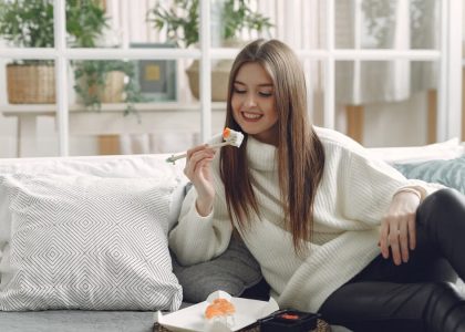 Smiling young lady in warm knitted sweater enjoying fresh sushi on wicker tray while relaxing on cozy sofa with soft cushions in modern living room